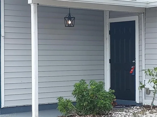 Entryway featuring a dark paneled door, light gray siding, an overhead geometric pendant light, a concrete porch, and established shrubbery