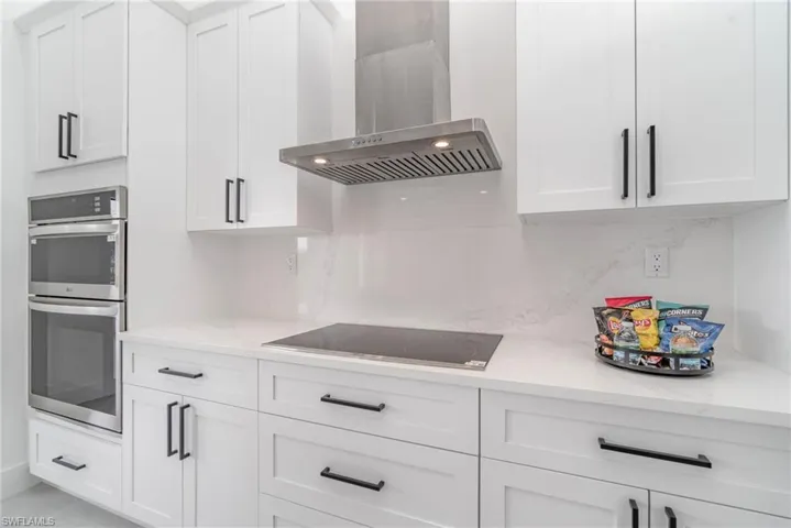 Kitchen with light stone counters, black electric cooktop, white cabinetry, wall chimney exhaust hood, and double oven