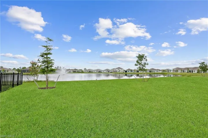 Expansive green lawn with mature trees and a black metal fence, overlooking a substantial body of water with a fountain feature