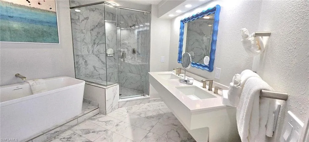 Full bath with double vanity, a freestanding bath, a marble finish shower, a textured wall, and light marble finish flooring