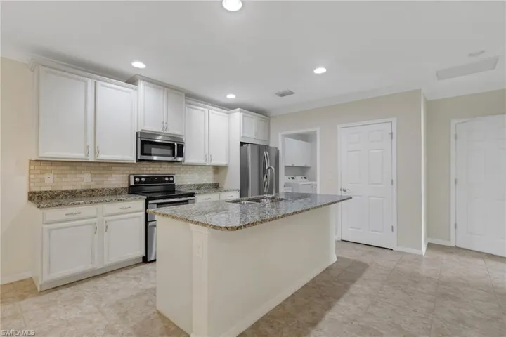 Kitchen featuring appliances with stainless steel finishes, light stone countertops, an island with sink, white cabinetry, and decorative backsplash