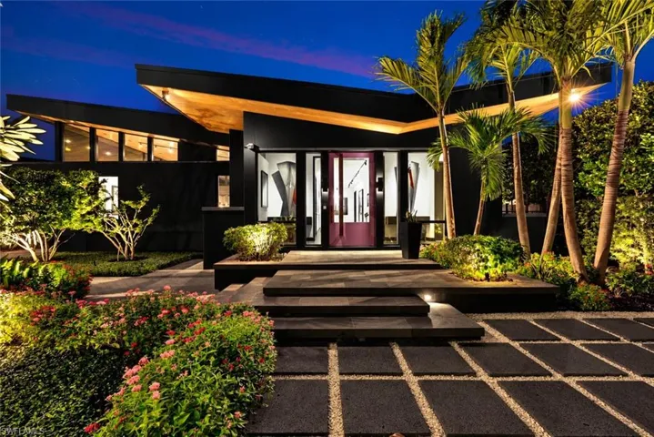 Modern architectural design featuring a prominent angular roofline, a striking red entry door, a multi-tiered entry staircase, and extensive landscaping with palm trees