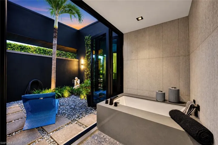 Freestanding soaking tub with matte black fixtures, textured wall tiling, recessed lighting, and direct access to an enclosed outdoor garden space featuring a palm tree and stone landscaping