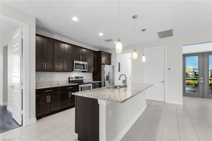 Kitchen with dark brown cabinets, stainless steel appliances, pendant lighting, a center island with sink, and light stone counters
