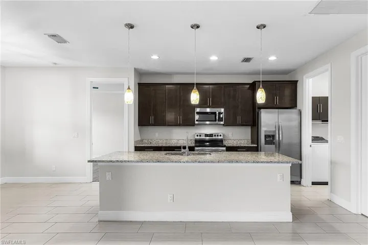 Kitchen with dark brown cabinetry, light stone counters, stainless steel appliances, decorative light fixtures, and an island with sink