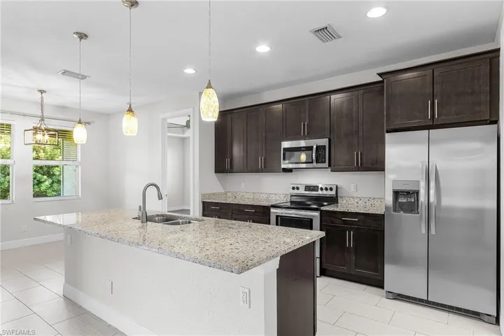 Kitchen featuring appliances with stainless steel finishes, dark brown cabinets, decorative light fixtures, light stone countertops, and a kitchen island with sink