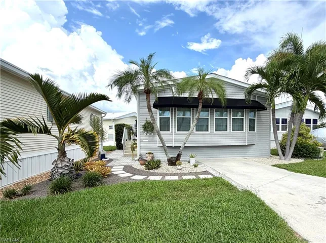 Exterior featuring light gray siding, multiple palm trees, a well-maintained lawn, a concrete driveway, and a decorative stone pathway