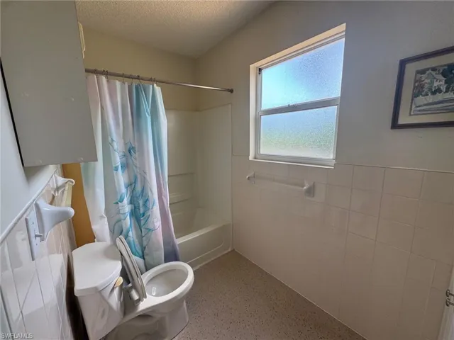 Bathroom featuring tile walls, a wainscoted wall, shower / tub combo, a textured ceiling, and dark aggregate flooring
