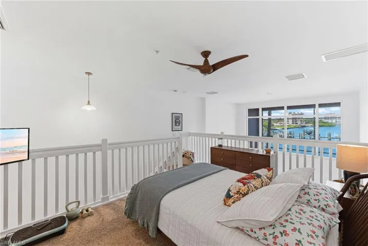 Loft space featuring a white baluster railing, a large window bank with water scenery, and a ceiling fan with wood-finish blades