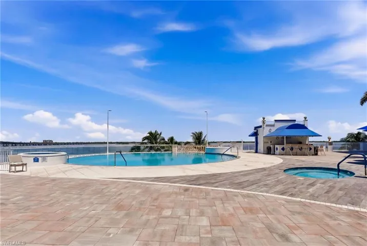 Expansive paver patio with a curved swimming pool, integrated spa, and an outdoor bar area under a blue canopy, all overlooking a body of water