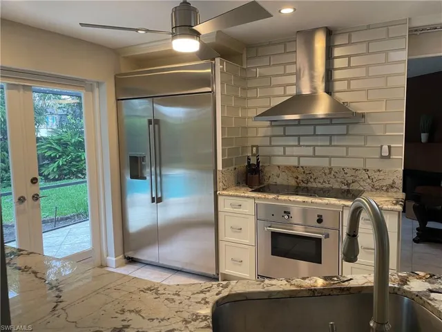 Kitchen with light stone counters, white cabinets, appliances with stainless steel finishes, wall chimney range hood, and decorative backsplash