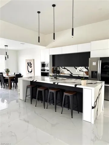 Kitchen featuring hanging light fixtures, a large island with sink, light marble finish floors, modern cabinets, and white cabinets