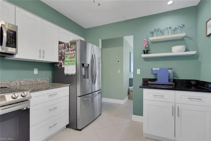 Kitchen featuring white shaker-style cabinetry, stainless steel appliances, contrasting stone-finish countertops, and wall-mounted shelving