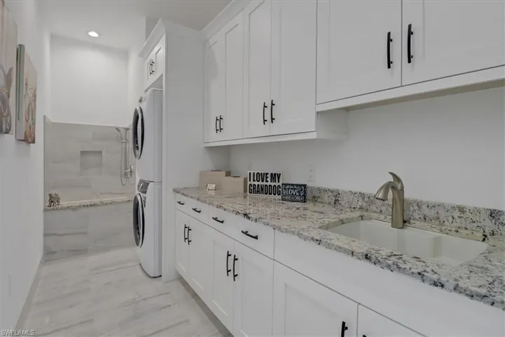 Laundry room featuring stacked washer/dryer, a custom Dog Shower, laundry sink, and cabinets above and below with granite counters