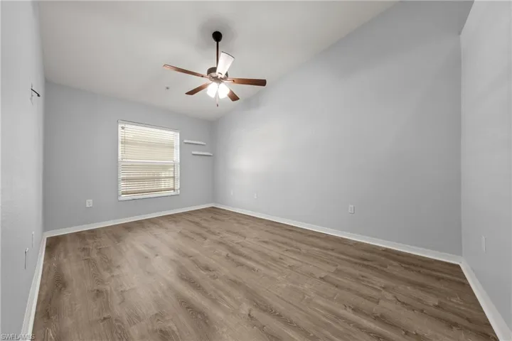 Spacious room featuring wood-finish flooring, a window with horizontal blinds, two wall-mounted shelves, and a ceiling fan with light fixture
