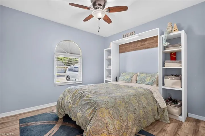 Built-in storage shelving, wood-finish flooring, arched window with blinds, ceiling fan, and wall-mounted headboard with integrated wood accent
