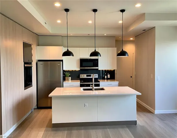 Modern kitchen featuring a central island with an integrated sink, white cabinetry, integrated stainless steel appliances, pendant lighting, and wood-finish flooring