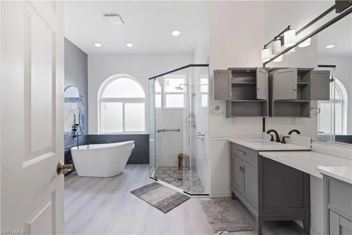 Contemporary bathroom featuring a freestanding tub, glass-enclosed shower, dual vanity with quartz countertops, gray cabinetry, and light wood-finish flooring