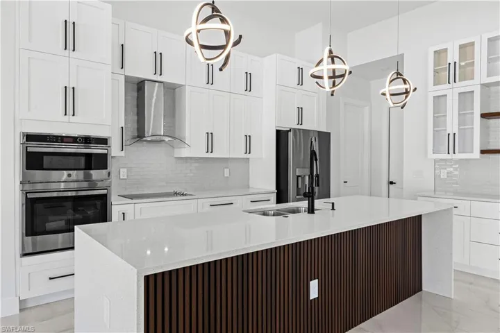 Kitchen featuring wall chimney exhaust hood, light stone counters, hanging light fixtures, appliances with stainless steel finishes, and white cabinets