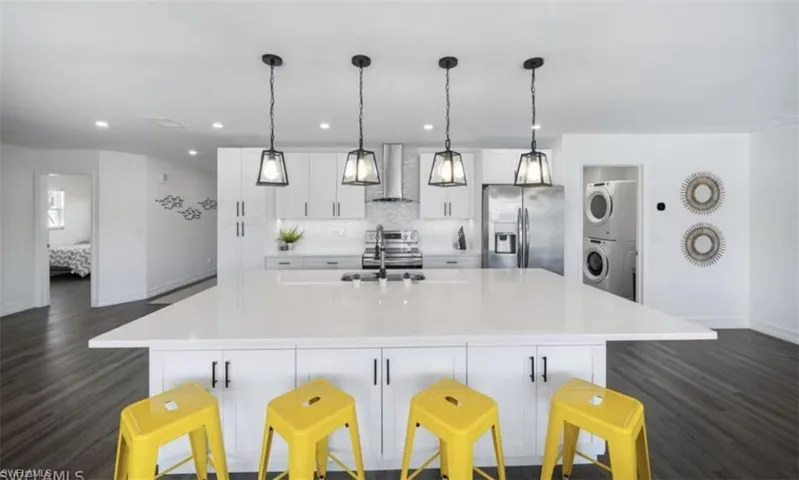 Kitchen featuring light countertops, appliances with stainless steel finishes, stacked washer and clothes dryer, wall chimney exhaust hood, and a large island with sink