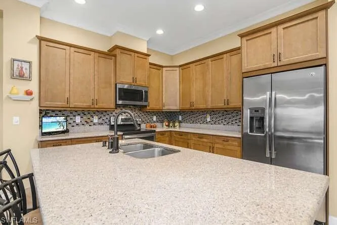 Kitchen featuring an island with sink, stainless steel appliances, wood finish cabinets, decorative backsplash, and light stone countertops