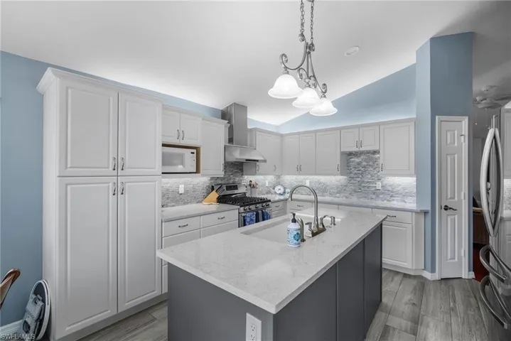 Kitchen featuring wall chimney range hood, hanging light fixtures, white cabinetry, stainless steel appliances, and a center island with sink