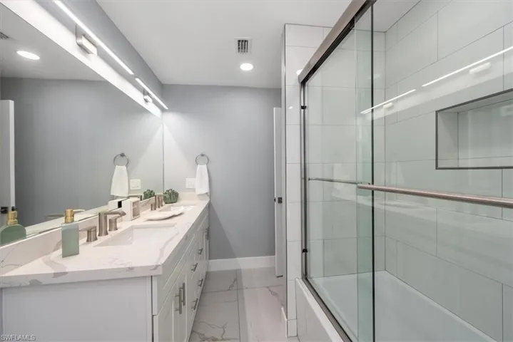 Bathroom featuring marble look tile flooring, baseboards, bath / shower combo with glass door, double vanity, and recessed lighting
