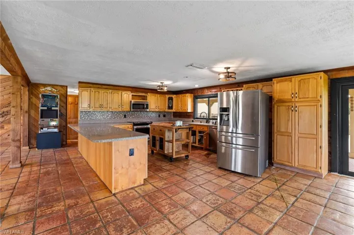 Spacious kitchen featuring extensive natural wood cabinetry, stainless steel appliances, a large central island with a stone-finish countertop, a mosaic tile backsplash, and terracotta-style floor tiles