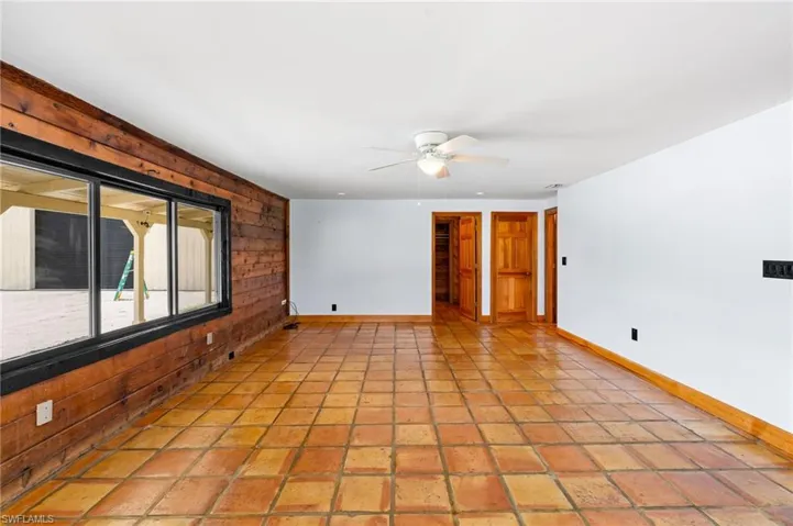 Spacious room featuring terra cotta tile flooring, a wide window with a black frame, and wood paneling on one wall