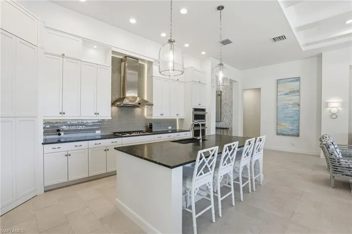 Kitchen featuring a kitchen island with sink, wall chimney range hood, decorative light fixtures, white cabinetry, and backsplash