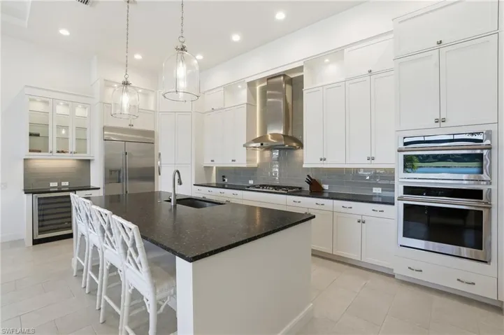 Kitchen with glass insert cabinets, a kitchen island with sink, white cabinetry, wall chimney exhaust hood, and appliances with stainless steel finishes