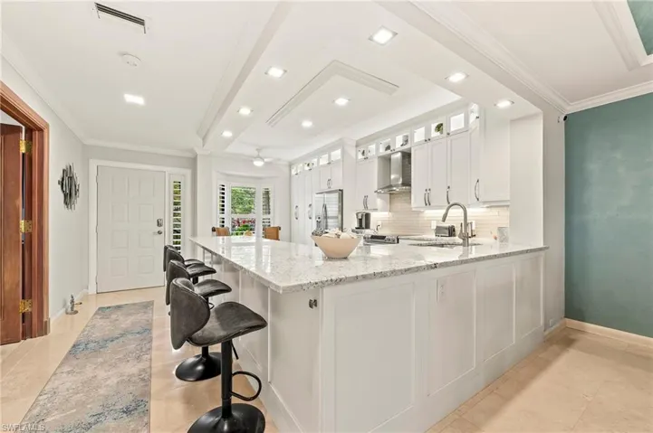 Kitchen featuring white cabinetry, kitchen peninsula, a breakfast bar area, wall chimney range hood, and light stone countertops