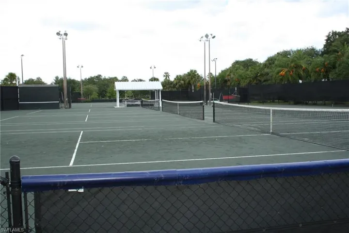 Tennis courts featuring a hard court surface, permanent nets, tall light poles, and a shaded seating area