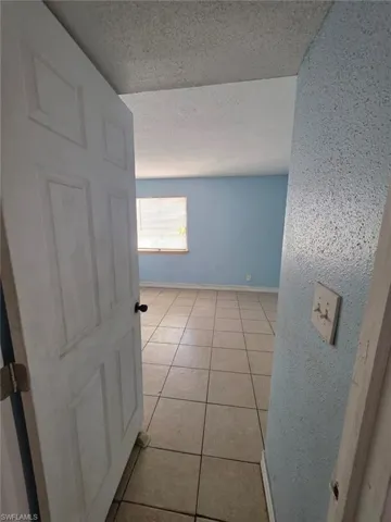 Spacious interior room featuring light blue walls, neutral-toned square tile flooring, a window with horizontal blinds, and a textured ceiling