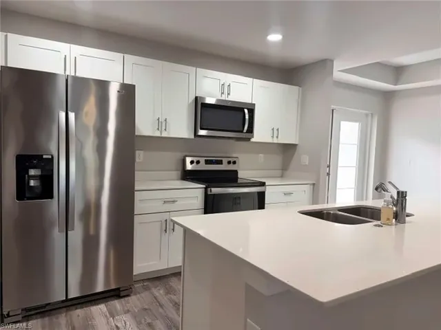 White shaker cabinetry, stainless steel appliances, light-toned countertops, wood-finish flooring, and a double basin sink