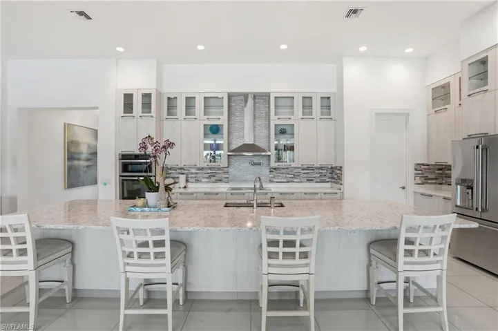 Kitchen with glass insert cabinets, appliances with stainless steel finishes, wall chimney range hood, light stone countertops, and a kitchen breakfast bar