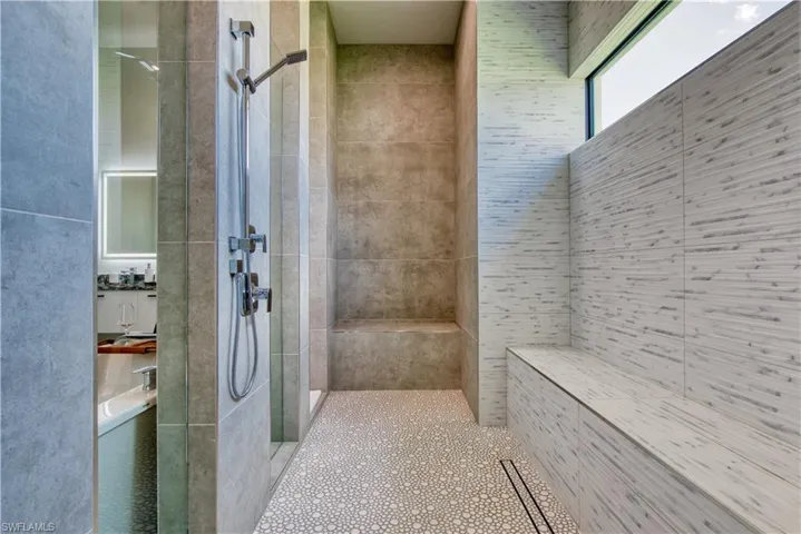 Walk-in shower with textured pebble-finish flooring, two shower heads, integrated bench seating, linear drainage system, and a high-set horizontal window