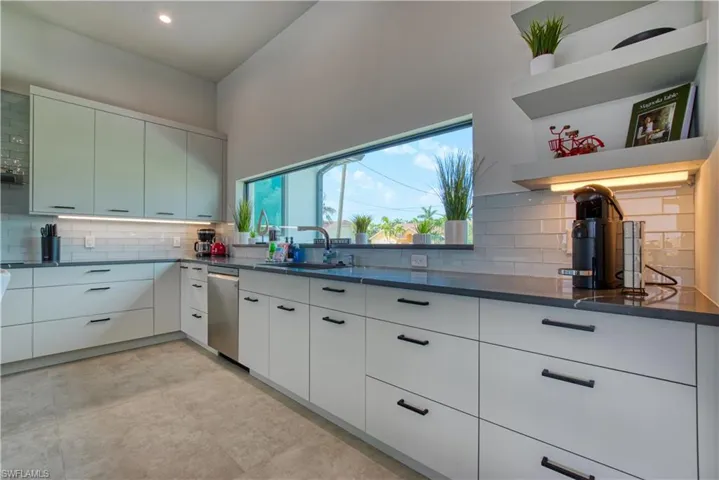 Contemporary kitchen featuring a wide picture window, vaulted ceiling, light-toned flat-panel cabinetry with black hardware, dark countertops, and a subway tile backsplash