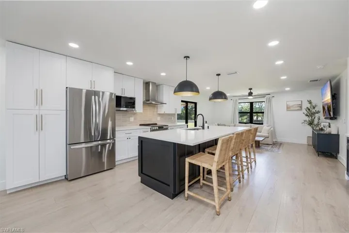 Kitchen with appliances with stainless steel finishes, decorative light fixtures, white cabinetry, a center island with sink, and wall chimney exhaust hood