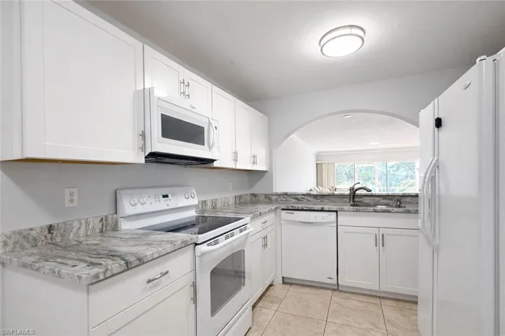 Kitchen with white appliances, white cabinets, light tile patterned floors, arched walkways, and light stone countertops