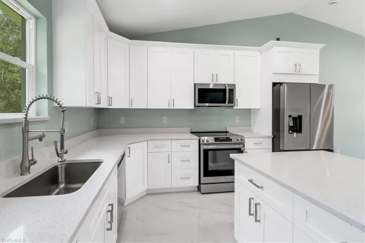 Kitchen featuring white cabinets, sink, appliances with stainless steel finishes, light stone countertops, and vaulted ceiling
