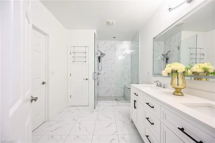 Spacious bathroom featuring a double vanity with ample storage, a large mirror, a glass-enclosed shower with a built-in bench, and white tile flooring with veining