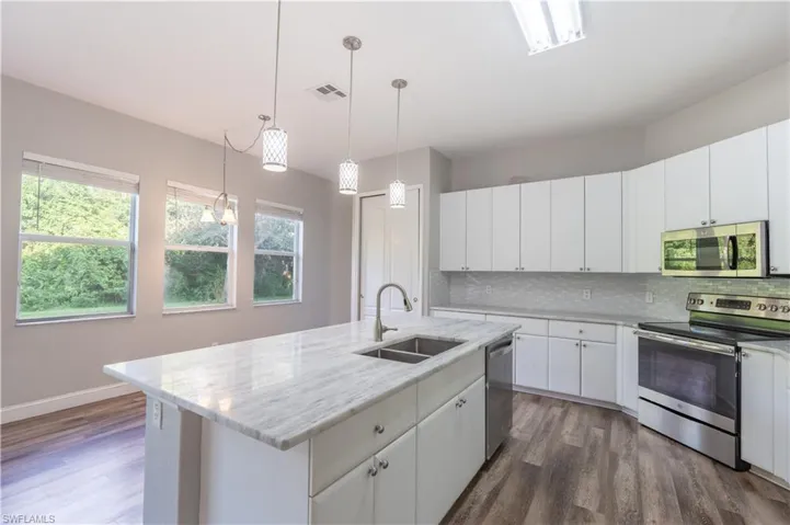 Kitchen featuring appliances with stainless steel finishes, backsplash, white cabinetry, decorative light fixtures, and a center island with sink