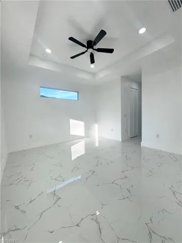 Empty room with a raised ceiling, light marble finish flooring, recessed lighting, and ceiling fan