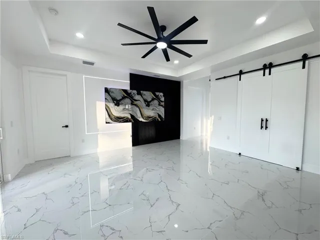 Unfurnished bedroom featuring a barn door, a tray ceiling, a ceiling fan, light marble finish floors, and recessed lighting