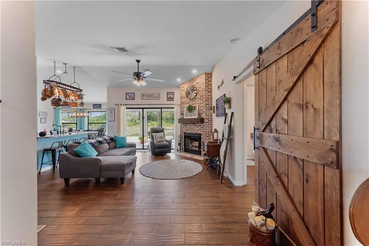 Living area with a barn door, ceiling fan with notable chandelier, dark wood-type flooring, visible vents, and a brick fireplace