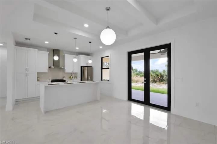 Kitchen featuring stainless steel fridge, wall chimney range hood, decorative light fixtures, white cabinets, and an island with sink