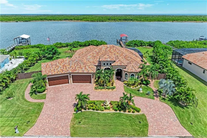 Waterfront property featuring a red tile roof, curved paver driveway, lush landscaping, and a private dock with a covered boat lift