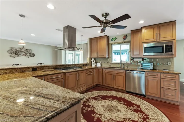 Kitchen featuring dark wood-type flooring, stainless steel appliances, island exhaust hood, stone countertops, and ceiling fan