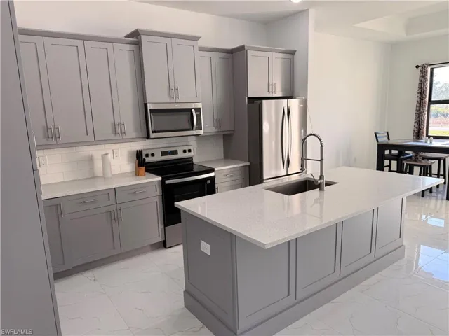 Kitchen with gray cabinetry, stainless steel appliances, light stone countertops, tasteful backsplash, and an island with sink